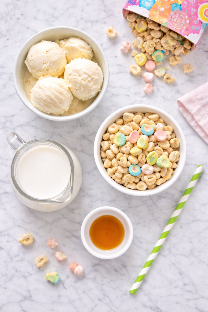 Flat lay of Lucky Charms milkshake ingredients including vanilla ice cream, milk, cereal, and vanilla extract on a white marble countertop