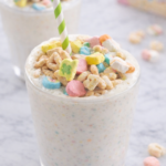 Lucky Charms milkshake with colorful marshmallow specks in a simple glass on a white marble countertop with green and white striped paper straw