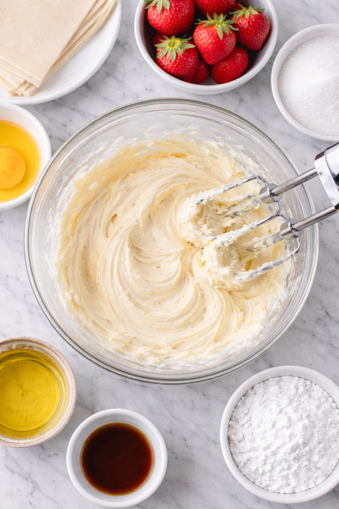cream cheese, sugar, and vanilla being mixed into cheesecake filling in a glass bowl on a marble counter