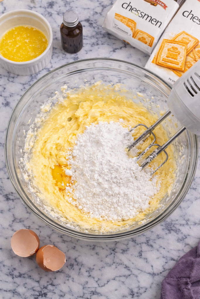 Cream cheese, eggs, powdered sugar, butter, and vanilla being mixed in a glass bowl to make chessman pie filling on a marble countertop