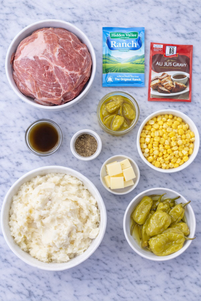 Flat lay of ingredients for Mississippi pork roast mashed potato bowls including pork roast, Hidden Valley ranch packet, au jus mix, pepperoncini, butter, corn and mashed potatoes on marble counter