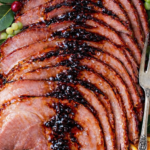 Sliced glazed ham on a wooden cutting board with orange slices, green grapes, bay leaves, and curly kale garnish