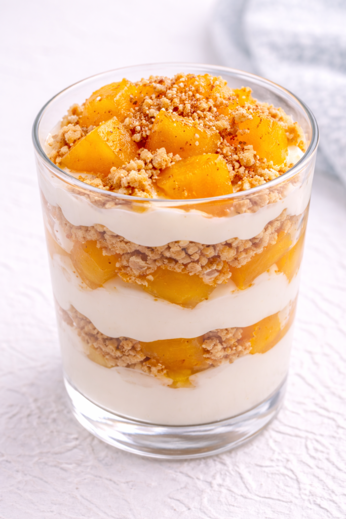 Peach pudding parfait layered with graham cracker crumbs and vanilla pudding in a plain glass