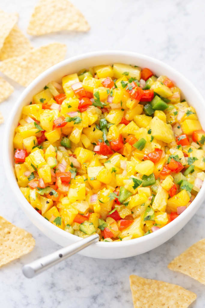 Bright pepper mango salsa in a white bowl on a marble counter with tortilla chips around it
