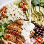 Grilled chicken rice bowl with avocado, black beans, pico de gallo, and creamy garlic cilantro sauce being poured over the top