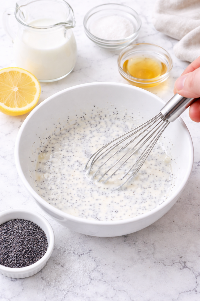 Whisking creamy poppyseed dressing in a white bowl on a marble countertop with simple ingredients nearby