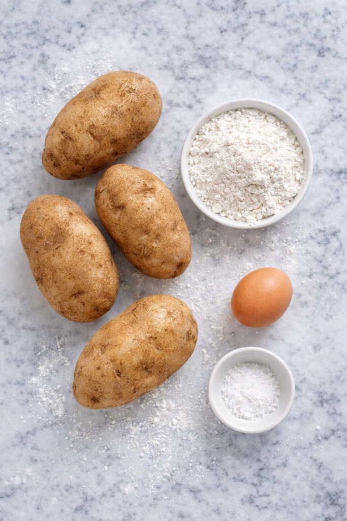 Flat lay of potato gnocchi ingredients including russet potatoes, flour, egg, and salt on a white marble countertop