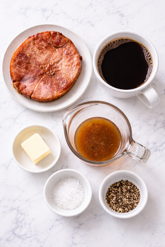 Flat lay of ingredients for red eye gravy including ham steak, black coffee, butter, and ham drippings on a marble countertop