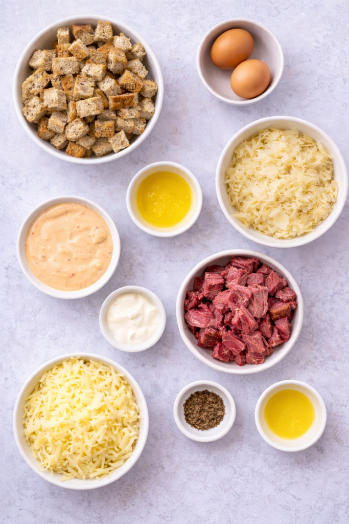 Flat lay of Reuben casserole ingredients including corned beef, sauerkraut, Swiss cheese, rye bread cubes, eggs, Thousand Island dressing, sour cream, butter, and caraway seeds in white bowls.