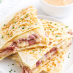 Reuben quesadilla stacked on a white round plate, filled with corned beef, Swiss cheese, and sauerkraut, topped with fresh chives and served with creamy dressing.