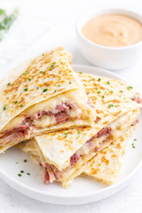 Reuben quesadilla stacked on a white round plate, filled with corned beef, Swiss cheese, and sauerkraut, topped with fresh chives and served with creamy dressing.