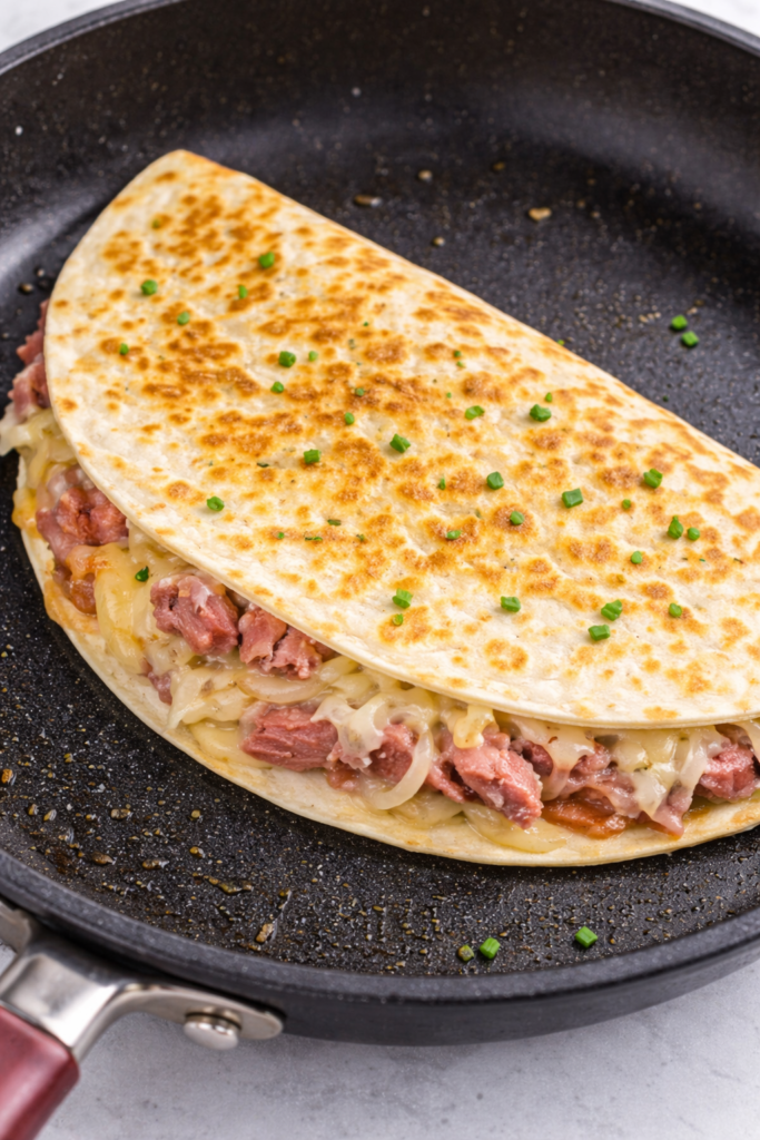 Reuben quesadilla cooking in a skillet with corned beef, sauerkraut, and melted Swiss cheese inside a golden crispy tortilla topped with fresh chives.