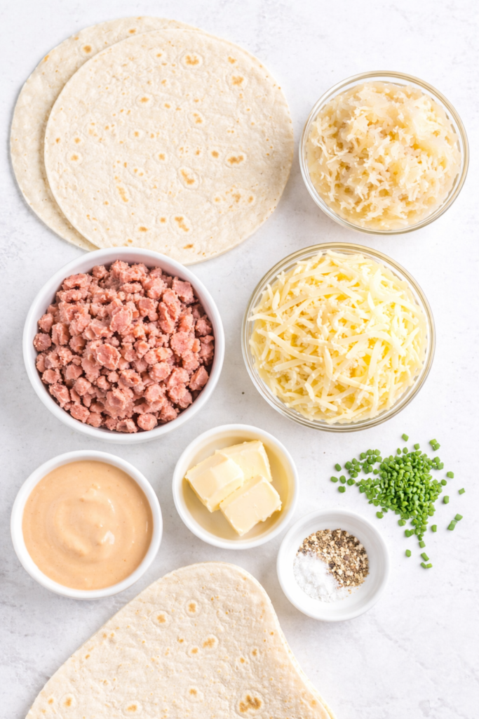 Flat lay of Reuben quesadilla ingredients including flour tortillas, corned beef, sauerkraut, shredded Swiss cheese, Russian dressing, butter, chives, salt, and pepper on a marble countertop.