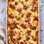Bird’s eye view of cheesy Reuben style casserole with corned beef, sauerkraut, and Swiss cheese baked until bubbly in a white dish with wooden serving spoon.