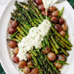 Roasted asparagus and pearl onions on a white platter with creamy herb Boursin sauce being poured over the vegetables on a light marble background.