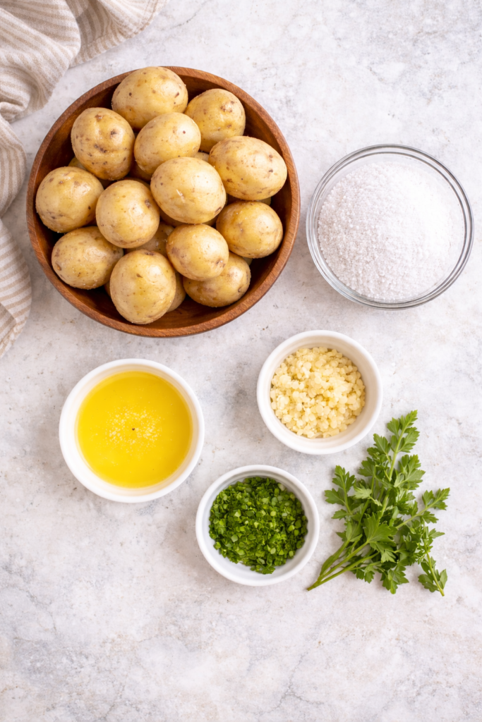 Flat lay of ingredients for salt potatoes including baby potatoes, coarse salt, butter, garlic, and parsley on a marble countertop