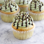 Shamrock shake cupcakes with mint buttercream, chocolate syrup drizzle, and chopped Andes mints on a marble countertop.