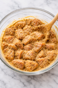 Chicken cubes marinating in yogurt and shawarma seasoning in a glass bowl on a marble countertop