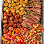 Sheet pan Brazilian steakhouse dinner with sliced steak, spicy sausage, roasted potatoes, peppers, onions and fresh tomato onion vinaigrette on a marble countertop.