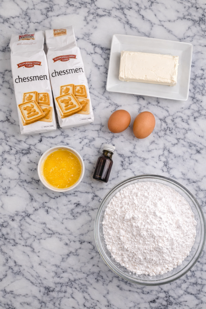 Flat lay of ingredients for sheet pan chessman pie including chessman cookies, cream cheese, eggs, butter, vanilla, and powdered sugar on a marble counter
