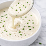 Creamy shortcut hollandaise sauce made with mayonnaise, lemon juice, and milk in a white bowl with black pepper and chopped chives on a marble countertop