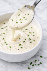 Creamy shortcut hollandaise sauce made with mayonnaise, lemon juice, and milk in a white bowl with black pepper and chopped chives on a marble countertop