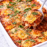Spoon scooping cheesy ravioli lasagna from a white baking dish with marinara sauce, melted mozzarella, torn basil, parmesan, and red pepper flakes on a marble countertop