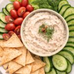 Smoked tuna dip in a bowl on a white platter with cucumbers, cherry tomatoes, and pita chips on a marble countertop