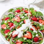 Spinach salad with strawberries, red onion, feta, and pecans on a white platter with creamy poppyseed dressing being drizzled over top on a marble counter