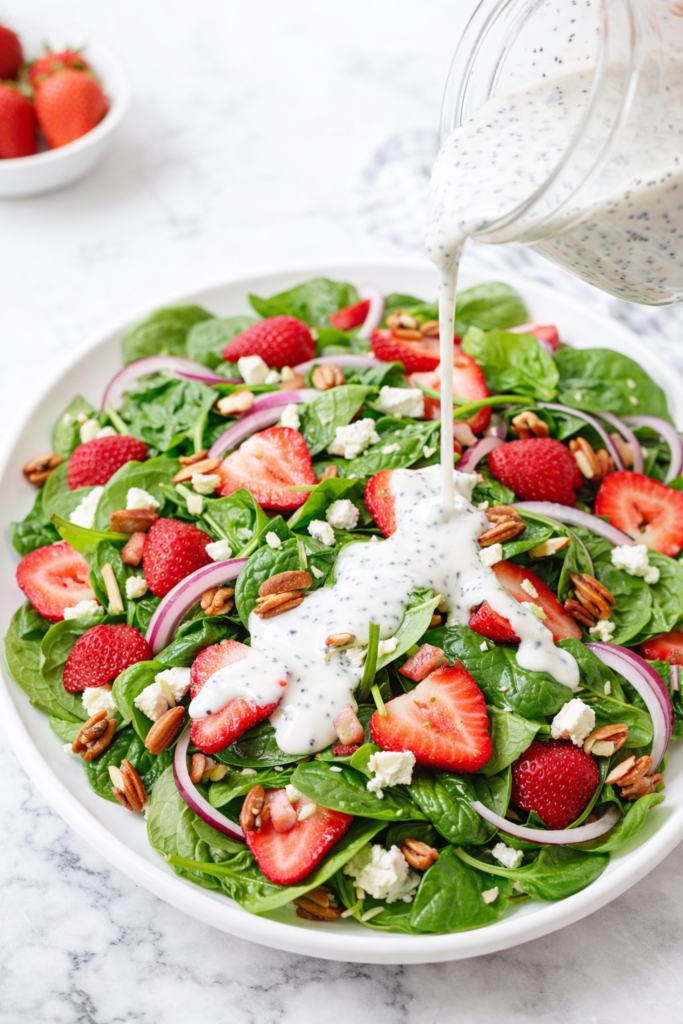 Spinach salad with strawberries, red onion, feta, and pecans on a white platter with creamy poppyseed dressing being drizzled over top on a marble counter