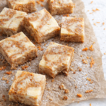 Swirled carrot cake cheesecake bars on wrinkled brown parchment paper with powdered sugar, shredded carrots, crumbs, and a bite taken out, styled on a marble countertop