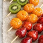 Tanghulu candied fruit skewers including strawberries, mandarin oranges, kiwi, and grapes on wrinkled parchment paper over a marble countertop