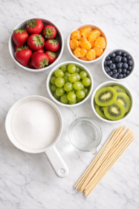Flat lay of ingredients for tanghulu including strawberries, grapes, kiwi, mandarin oranges, blueberries, sugar, water, and wooden skewers on a marble countertop