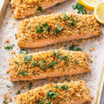 Walnut rosemary crusted salmon fillets baked on a parchment lined sheet pan with lemon wedges
