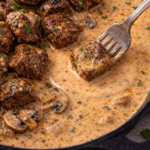 fork dipping whiskey marinated steak tip into creamy garlic paprika sauce with mushrooms and onions in cast iron skillet