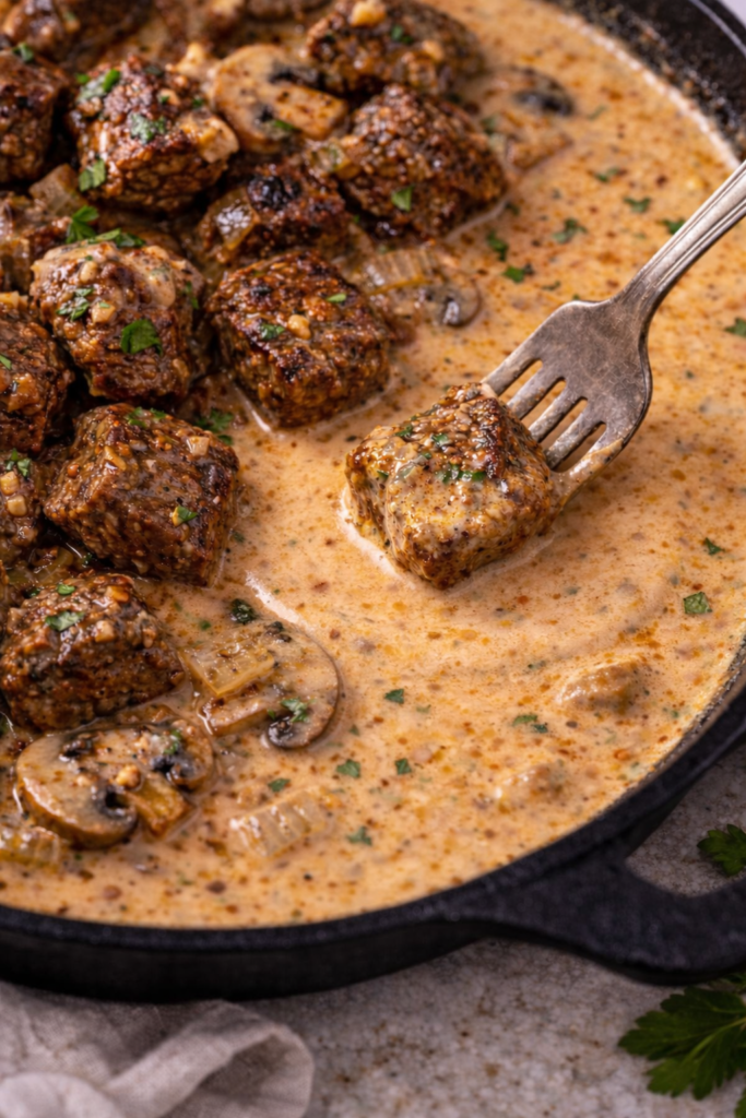 fork dipping whiskey marinated steak tip into creamy garlic paprika sauce with mushrooms and onions in cast iron skillet