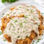 Crispy chicken fried steak smothered in creamy country gravy with black pepper and parsley on a white plate