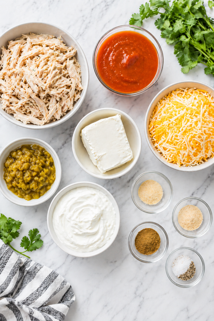 flat lay of chicken enchilada dip ingredients including shredded chicken enchilada sauce cream cheese sour cream shredded cheese and green chiles on a marble countertop