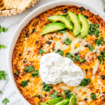 creamy chicken enchilada dip in a round white baking dish topped with melted cheese avocado and sour cream on a marble countertop