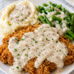Chicken fried steak with creamy country gravy served with mashed potatoes and green beans on a white plate