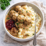 Copycat IKEA Swedish meatballs over creamy mashed potatoes with rich gravy and lingonberry jam in a white bowl on a marble countertop