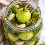 Fork piercing a whole pickled green tomato in a jar with garlic, dill, and spices on a marble countertop