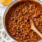 Homemade pork and beans in a white Dutch oven topped with fresh chives and served with cornbread muffins