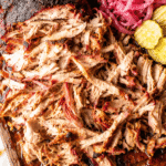 Smoked pork shoulder with crispy bark and juicy pulled pork on a dark wooden cutting board over a white marble countertop with barbecue sauce, pickles, pickled onions, and pork juices dripping onto the marble.