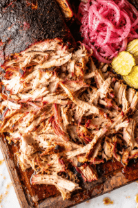 Smoked pork shoulder with crispy bark and juicy pulled pork on a dark wooden cutting board over a white marble countertop with barbecue sauce, pickles, pickled onions, and pork juices dripping onto the marble.