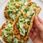 Golden crispy chicken Caesar smash tacos topped with fresh Caesar salad, shaved parmesan, and croutons on a white plate with a hand holding one taco folded open over a marble countertop.