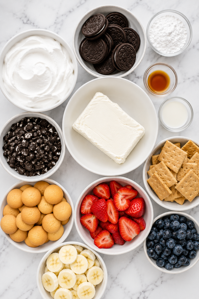 Flat lay of ingredients for cookies and cream cheesecake dip including cream cheese, whipped topping, chocolate sandwich cookies, powdered sugar, vanilla, strawberries, blueberries, banana slices, vanilla wafers, and graham crackers on a white marble countertop.