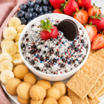 Creamy cookies and cream cheesecake dip in a simple white bowl topped with crushed chocolate cookies and fresh strawberries, surrounded by blueberries, banana slices, vanilla wafers, and graham crackers on a white marble countertop.