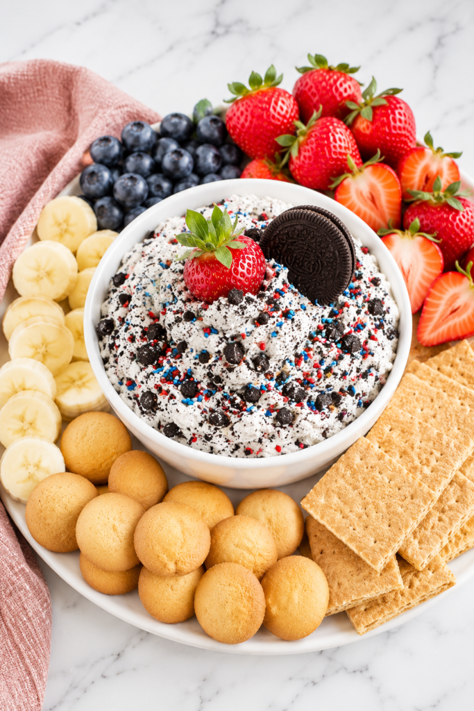 Creamy cookies and cream cheesecake dip in a simple white bowl topped with crushed chocolate cookies and fresh strawberries, surrounded by blueberries, banana slices, vanilla wafers, and graham crackers on a white marble countertop.