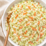 Creamy copycat KFC coleslaw in a white serving bowl with visible shredded carrots and celery seed garnish, styled on a white marble countertop with a neutral linen cloth and copper spoon.